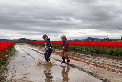 Boys jumping on puddle at road amidst field against cloudy sky