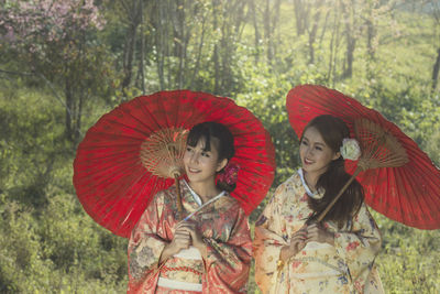 Smiling young women in kimono holding red umbrellas against trees