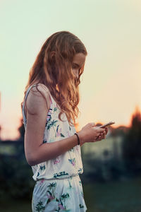 Side view of girl using mobile phone against sky during sunset