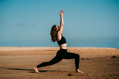 Slim female in sportswear standing on sandy beach in high lunge crescent variation pose and practicing yoga while looking up