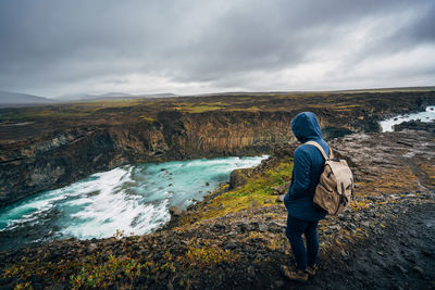 Rear view of person standing by pond against cloudy sky