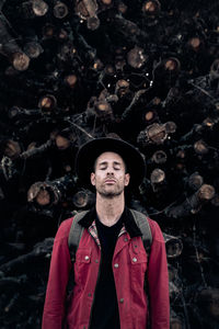 Tranquil adult man in hat and red shirt closing eyes against pile of logs in nature