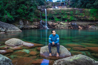 Young man isolated sitting on rock with pristine waterfall background at morning in forests