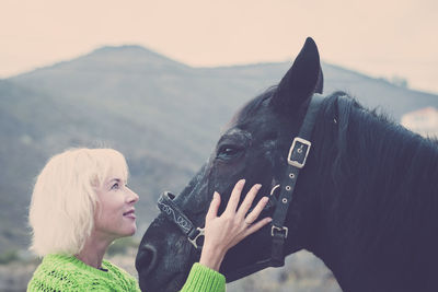 Side view of smiling woman stroking black horse against mountain