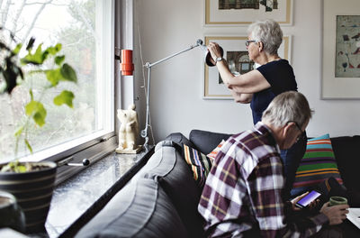 Senior woman adjusting floor lamp by man sitting on sofa in living room at home