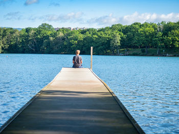 Blond hair boy sits on lake mole and looks at water, view from back. calm water of city park pond