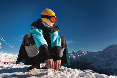 Young skier, wearing ski glasses in the mountains and colorful clothes, sitting over clear sky and