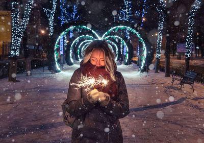 Woman holding illuminated sparklers on snowy footpath at night during christmas