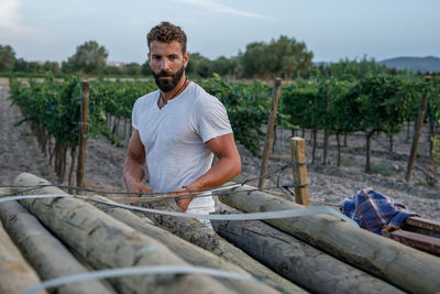 Bearded man in white t shirt putting heavy log into stack near grapevines during work on farm