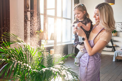 Young attractive woman with her toddler girl watering plants in home. making domestic work