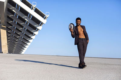 Portrait of man holding hat while standing on road against clear sky