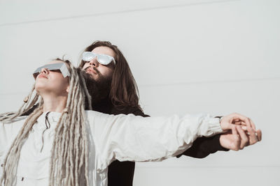 Young couple wearing sunglasses with arms outstretched standing against wall