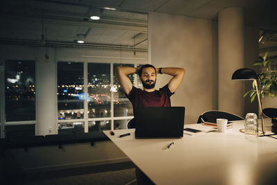 Tired male entrepreneur looking at laptop while sitting with hands behind head in creative office