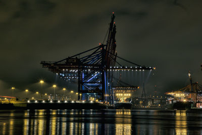 Illuminated cranes over sea against sky at night