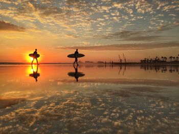 Silhouette surfers walking at beach against sky during sunset