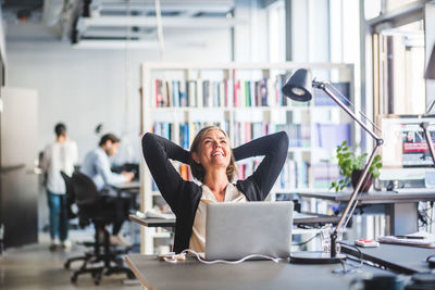 Mature businesswoman sitting with hands behind head at desk in office