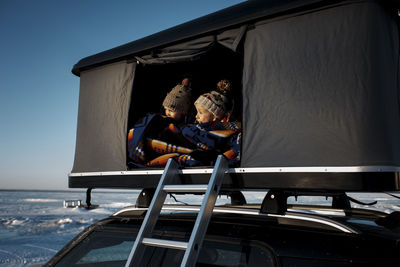 Siblings looking away while sitting in roof tent on car