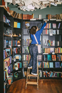 Full length of woman selecting books while standing on ladder near shelf in bookstore