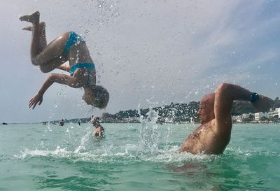 Girl backflipping with shirtless father in sea against sky