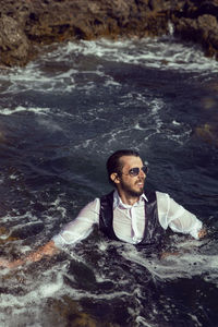 Man with a beard and sunglasses in clothes a vest and a white shirt swims in the sea among the rocks