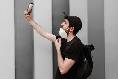 Side view of cheerful unshaven male in black wear with rucksack taking selfie on cellphone during quarantine period