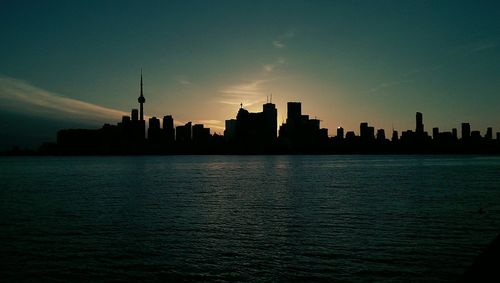 Silhouette buildings by river in city against sky during sunset