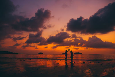 Silhouette couple with arms raised holding hands at beach against orange sky during sunset