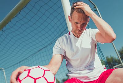 Low angle view of worried soccer player sitting by goal post against sky