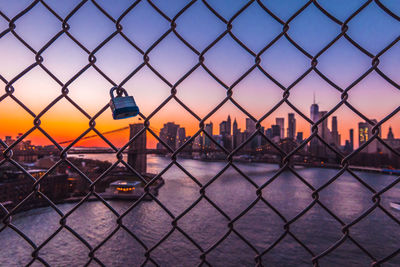 Lock hanging on chainlink fence over river in city during sunset