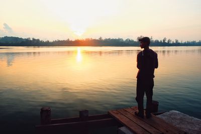 Man standing on jetty by lake against sky during sunset