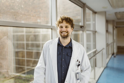 Portrait of smiling mature doctor wearing lab coat standing by window in hospital corridor