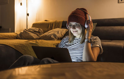 Smiling young woman wearing wireless headphones and using laptop in living room
