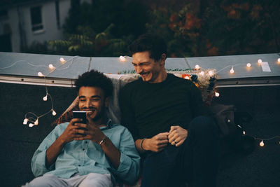 Smiling male friends looking at smart phone sitting on terrace during rooftop party in city