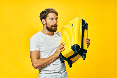 Young man holding suitcase against yellow background