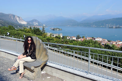 Portrait of woman sitting on railing against mountains and lake