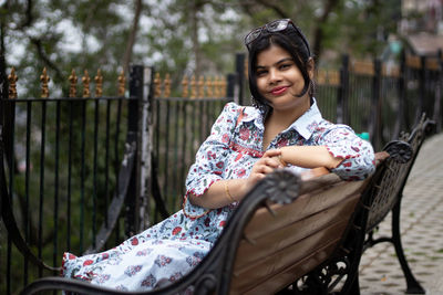 A pretty indian woman with sunglasses on head smiling and looking at camera while sitting on a bench