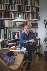 Serious mature therapist discussing with patient against bookshelf at home office