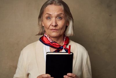 Smiling senior woman holding book standing against wall