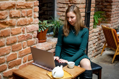 Young woman using phone while sitting on brick wall