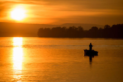 Silhouette man fishing on boat in lake against sky during sunset