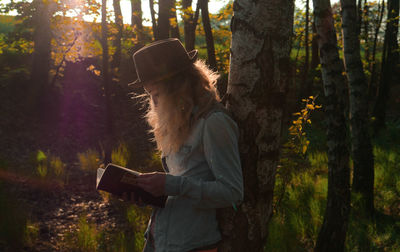 Side view of woman reading book by tree in forest