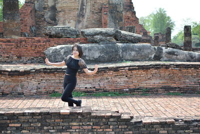 Portrait of young woman with arms outstretched standing on footpath against old ruin temple