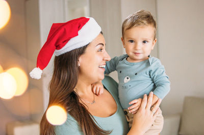 Mother wearing santa hat while carrying son at home