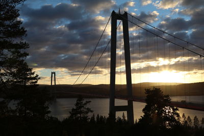 Silhouette of suspension bridge over river against cloudy sky