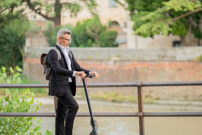 Side view full length young businessman in a suit riding an electric scooter while commuting to work 