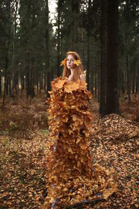 Thoughtful young woman covered with leaves standing at forest during autumn