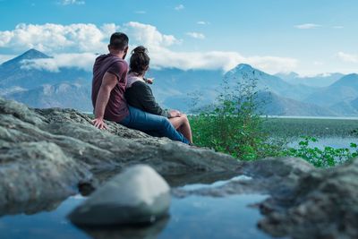 Friends sitting on rock by lake against sky