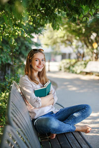 Portrait of happy young woman reading book while sitting on bench at park