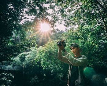 Young man photographing with camera while standing in forest during sunny day