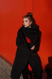 Fashionable teenage girl wearing black warm clothing standing against red wall during sunny day